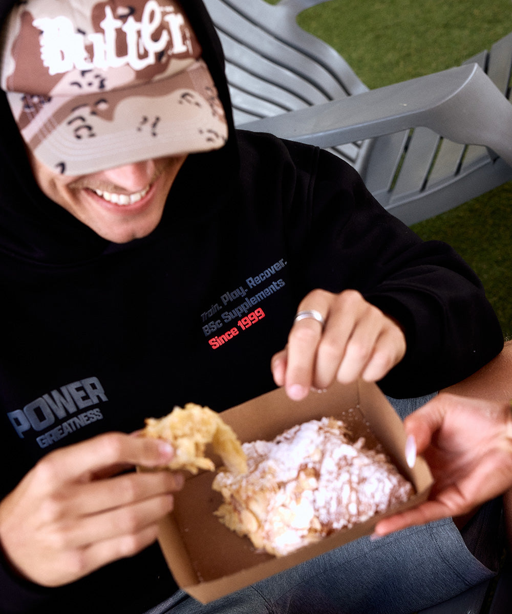 BSc Performance Hoodie in black, worn by a man eating a pastry on an outdoor chair